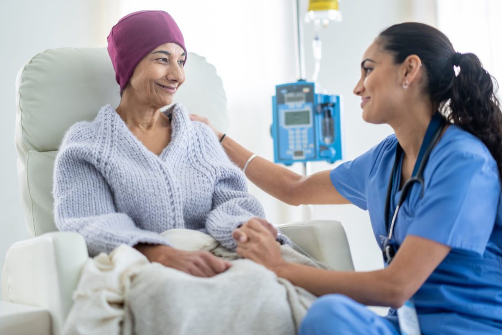 A senior woman battling cancer sits in a high back chair as she talks with her nurse during a Chemotherapy treatment. The patent is dressed casually in a sweater and has a blanket and headscarf on to keep her warm.